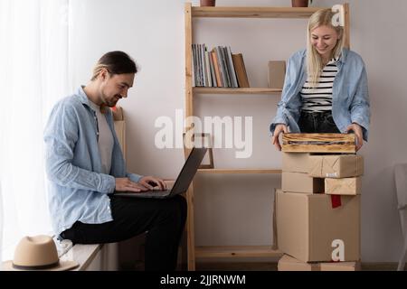 Junges Paar zieht in ein neues Zuhause. Glückliche Hausbesitzer, die mit einem Laptop in einer Wohnung sitzen. Eine Frau packt Dinge aus. Suche nach Hause dekorieren Ideen Stockfoto