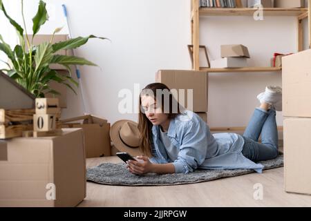 Junge kaukasische Frau benutzt Smartphone im Wohnzimmer des neuen Hauses mit Stapel von Kartons am Umzugstag, Frau schreibt während sie zu sich zieht Stockfoto