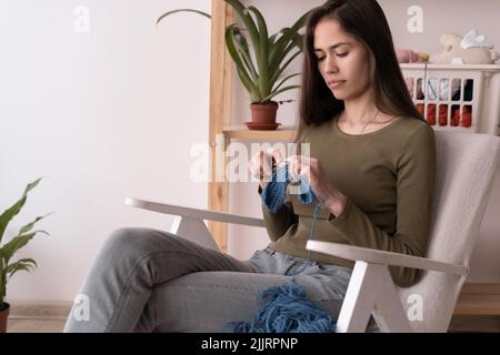 lateinische junge Frau strickt, während sie zu Hause auf einem Sessel sitzt, Hobby und Handarbeit, handgemachte Strickwaren, Freizeit und Hobby Stockfoto