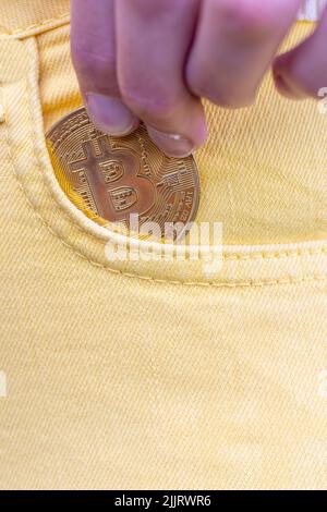 Nahaufnahme einer Frau, die Bitcoin in die gelbe Jeanstasche steckte, goldene Kryptowährung-Münze im Taschenhintergrund, Konzept der digitalen Währung virtuelles Geld. Hochwertige Fotos Stockfoto