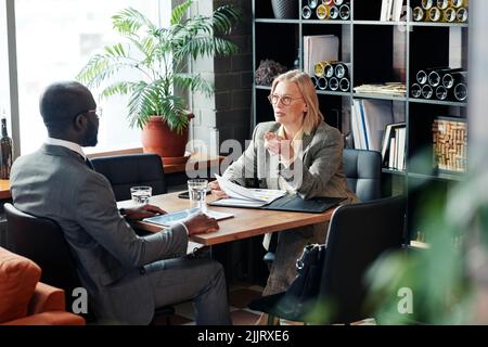 Reife Geschäftsfrau im Gespräch mit afrikanischen Geschäftsmann am Tisch mit Dokumenten, sie diskutieren Vertrag während der Sitzung im Restaurant Stockfoto