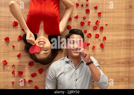 Direkt über dem Bild eines fröhlichen asiatischen Paares, das auf dem Boden liegt und ihre Augen mit Valentinskarten bedeckt, rote Rosenblätter, die überall verstreut sind Stockfoto