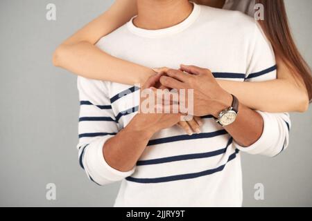 Nahaufnahme einer nicht erkennbaren Frau, die hinter ihrem Freund steht und ihn umarmt, grauer Hintergrund Stockfoto