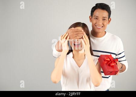 Hübscher asiatischer Mann mit gestreiftem Pullover, der seine Freundin angenehm überrascht: Er hält eine Geschenkbox und bedeckt ihre Augen mit Händen und grauem Hintergrund Stockfoto