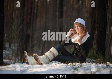 Ein kaukasisch blondes Mädchen, das einen weißen Winterhut und Schal trägt und im Schnee sitzt Stockfoto