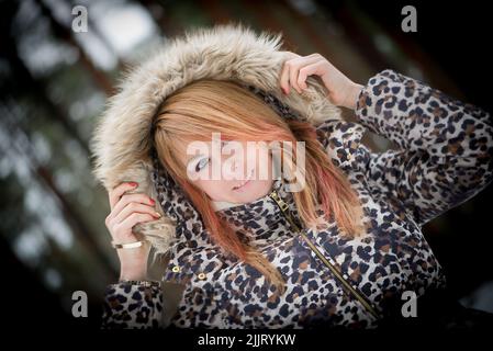 Ein Porträt einer kaukasischen jungen Frau, die einen Mantel mit Leopardenmuster im Winter in einem verschneiten Wald trägt Stockfoto