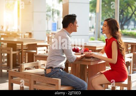 Profilansicht einer attraktiven jungen Frau im roten Kleid, die am Kaffeetisch im Freien sitzt, während ihr Freund ihre Hand zärtlich hält Stockfoto