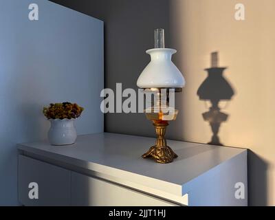 Schöne Vintage Öllampe mit Lampenschirm aus weißem Glas auf einem weißen Schrank. Schatten der Lampe an der Wand. Stockfoto