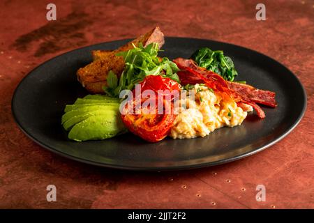 Ein Teller englisches Frühstück mit gebratenem Speck, Rührei, Toast, Avocado-Scheiben und Gemüse Stockfoto