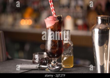 Eine Nahaufnahme von Cocktail aus schwarzer Kirsche mit Stroh neben Glasflasche und Löffel auf dem Tisch Stockfoto