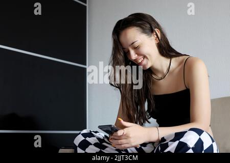 Hübsches junges Mädchen mit langen Haaren lachend, während sie mit dem Smartphone auf dem Sofa sitzt. Online-Kommunikation, positive Emotionen, Humor-Video ansehen Stockfoto