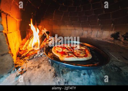 Ein schwarzes Tablett mit einer rohen Pizza mit Fleisch und Käse im Ofen in der Nähe eines Holzfeuers Stockfoto