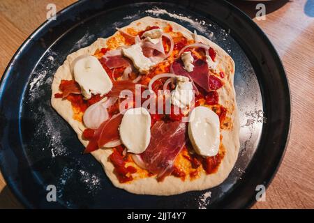 Eine Draufsicht auf eine rohe Pizza mit Fleisch und Käse auf einem schwarzen Tablett Stockfoto