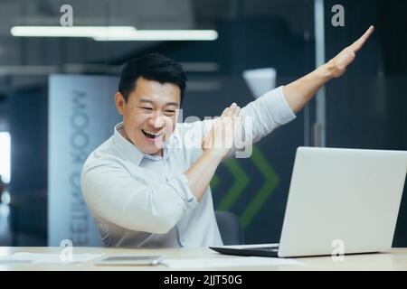 Erfolgreicher und gutaussehender junger Geschäftsmann, asiatischer Mann, der mit Händen tanzt und Kameraposen zeigt und lächelt. Sitzen am Schreibtisch im Büro, feiern Sieg, Erfolg, glücklich. Stockfoto