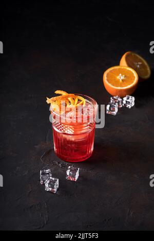 Negroni Cocktail mit Orange Twist und Pin, auf dunklem Hintergrund, und daneben Orange und Eis. Getränkefotografie, Kopierbereich. Selektiver Fokus. Stockfoto