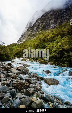 Eine vertikale Aufnahme eines nebligen Himmels, der eine felsige Klippe neben einem hellblauen Strom in den Milford Sounds bedeckt Stockfoto