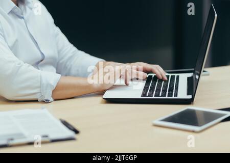 Nahaufnahme. Die Hände eines jungen Geschäftsmannes, eines Freiberuflers, eines IT-Mitarbeiters in einem weißen Hemd, der hinter einem Laptop arbeitet, auf einer Tastatur tippt, an einem Schreibtisch in einem modernen Büro. Stockfoto