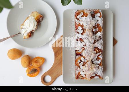 Rechteckiger appetitlich hausgemachter Aprikosenkuchen auf einem rechteckigen Teller, auf einem hölzernen Schneidebrett. Daneben ist ein runder Teller mit einem Stück Kuchen und einem Stockfoto