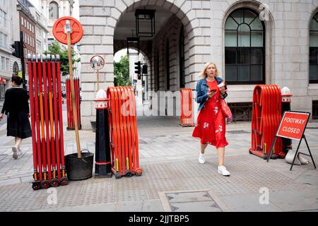 Fußgänger gehen zwischen den auf dem Bürgersteig befindlichen Ziehharmonika-Turtle-Gates, die von Banksmen während der Bauarbeiten in der City of London, dem Finanzdistrikt der Hauptstadt, am 27.. Juli 2022 in London, England, entfaltet werden können. Stockfoto