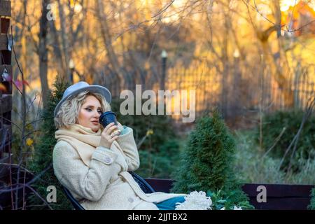 Eine Frau in einem beigen Hut und Mantel trinkt Kaffee in einem Café vor dem Hintergrund einer herbstlichen Landschaft. Reflexion, Kontemplation. Einsamkeit. Traurigkeit. Stockfoto