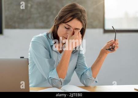 Überarbeitete Büroangestellte hält eine Brille in den Händen und massiert den Nasenrücken Stockfoto