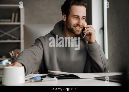 Junger Bartmann, der im Büro Kaffee trinkt und am Handy telefoniert Stockfoto
