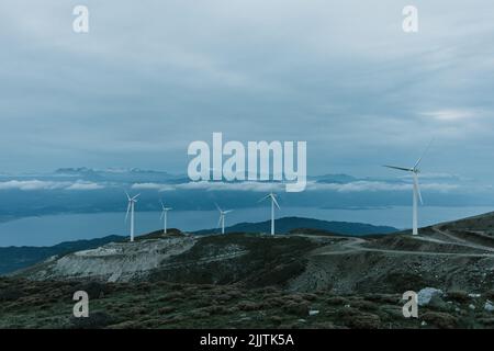 Die Windturbinen auf Hügeln am Seeufer gegen den blau bewölkten Himmel in Griechenland Stockfoto