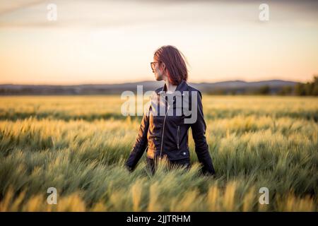 Ein schönes Porträt eines jungen Mädchens, das unter langem grünen Gras auf dem Feld gegen den Abendhimmel bei Sonnenuntergang steht Stockfoto