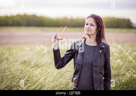 Eine schöne junge kaukasische Frau, die ihre Sonnenbrille hält, während sie an einem sonnigen Tag zwischen langem grünen Gras auf dem Feld steht Stockfoto