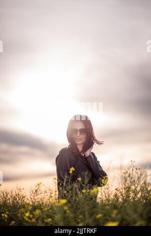 Eine schöne junge kaukasische Frau, die auf einem Feld mit blühenden gelben Blumen gegen den Abendhimmel bei Sonnenuntergang steht Stockfoto