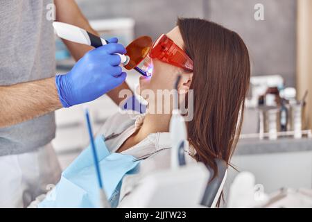 Professionelle männliche Arzt verwendet ultraviolette Lampe nach der Herstellung einer Füllung auf einem Zahn für freudige glückliche kaukasische Frau in modernen Zahnschrank Stockfoto