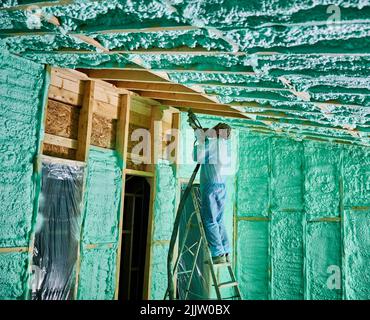 Männlich Baumeister isolierenden Holzrahmen Haus. Mann Arbeiter sprühen Polyurethan-Schaum in der zukünftigen Hütte, stehend auf der Leiter, mit Plural Komponente Pistole. Bau- und Isolierkonzept. Stockfoto