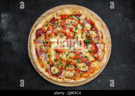 Pizza mit Salami, Schinken, Tomaten und würziger Sauce, auf einem runden Holzbrett auf grauem Beton. Draufsicht Stockfoto