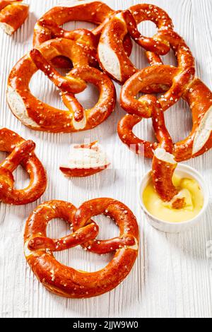 Weiche Brezeln in Form von Knoten gebacken und mit Salz auf weißem Holztisch mit Käsesauce bestreut, vertikale Ansicht von oben, Nahaufnahme Stockfoto