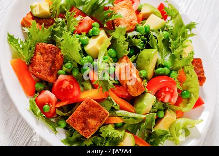 Nahaufnahme von Tofu-Salat mit Avocado, knackigen Salatblättern, grünen Erbsen, Tomaten, gelbem Paprika in einer weißen Schüssel Stockfoto