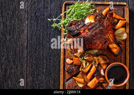 Knuspriger deutscher Schweinsschenkel, gebratener Schinkenbock, Schweinshaxe, mit Biersoße und Gemüse auf unhöflichem Holzbrett auf dunklem Holztisch, horizontal Stockfoto