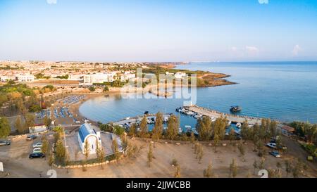 Luftbild des Sonnenuntergangs an der Küste und die weiße, gewaschene Kapelle am Strand Agia Triada, Protaras, Famagusta, Zypern von oben. Vogelperspektive auf Tour Stockfoto