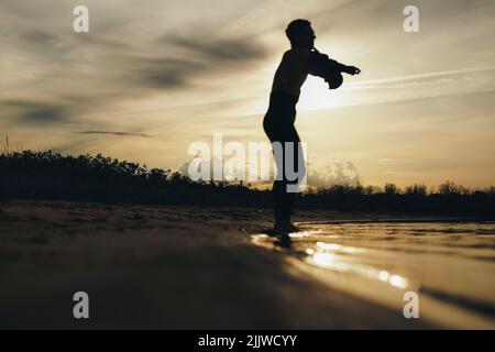 Silhouette eines männlichen Tauchers, der einen Neoprenanzug ansetzt, während er bei Sonnenuntergang am Strand steht. Abenteuerlicher junger Mann, der sich vorbereitet, um durch t ins Wasser zu kommen Stockfoto