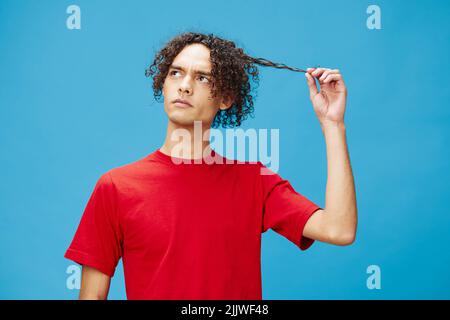 Nachdenklich nachdenklich unglücklich junge lockig gebräunt kaukasischen Kerl in grundlegenden roten T-Shirt posiert isoliert auf blauem Hintergrund. Lifestyle und Emotionen Stockfoto