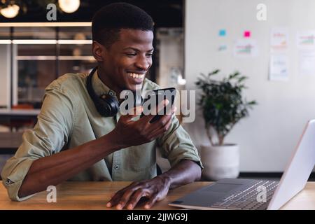 Lächelnder afroamerikanischer junger Geschäftsmann, der im Büro über einen Smartphone-Lautsprecher spricht Stockfoto