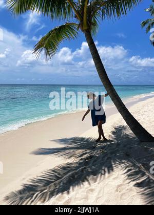 Junge Frau in blauem Kleid, die eine Palme hält, während sie auf einer tropischen Insel steht Stockfoto