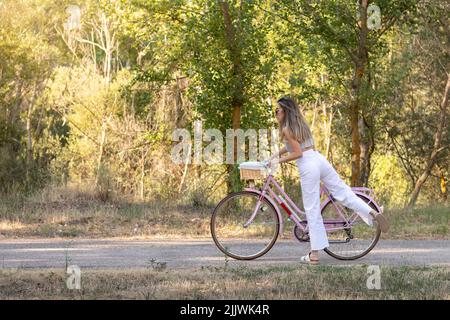 Seitenansicht der jungen eleganten Frau, die im Frühling auf einem rosafarbenen Retro-Fahrrad mitten in der Natur unterwegs ist Stockfoto