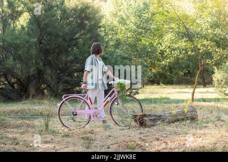 Seitenansicht eines nicht erkennbaren, hinten aussehenden jungen langhaarigen Mannes, der ein pinkes Vintage-Fahrrad im Park hält Stockfoto