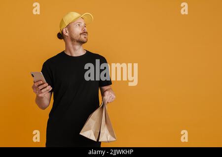 Weißer Lieferer mit Papiertüten, der sein Mobiltelefon auf gelbem Hintergrund isoliert hat Stockfoto