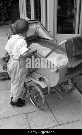 1960s, historisch, draußen auf einer Terrasse, ein kleiner Junge in Baumwoll-Latzhose, der an einem Kinderwagen der Zeit auf den Zehen steht, und ein Baby sieht, das darin liegt, bedeckt mit einer Decke, England, Großbritannien. Stockfoto
