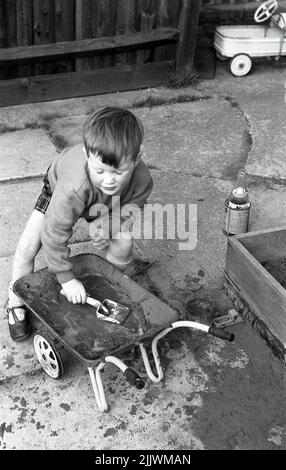 1960s, historisch, draußen auf einer Terrasse, ein kleiner Junge, der den Slushie herunterstachelt, nasser Sand in seiner Schubkarre, den er aus einem Sandkasten, England, Großbritannien, verlegt hat. Stockfoto