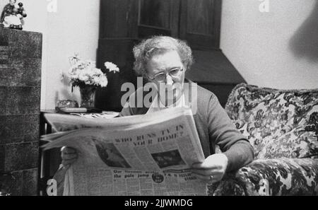 1960s, historisch, sitzend in einem gemustert Lounge Chair, eine eldelry Dame, die eine Zeitung liest, Schlagzeile, 'Wer läuft Fernsehen in Ihrem Haus?', England, Großbritannien. Stockfoto