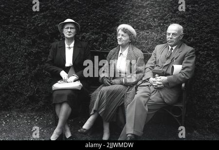 1960s, historisch, drei gut gekleidete ältere Menschen, die auf einer Parkbank sitzen, England, Großbritannien. Die Damen sind in eleganten Outfits, mit Handtaschen und Hüten, der Gentleman in Anzug und Krawatte, raucht eine Pfeife und hält eine Kamerahülle. Stockfoto