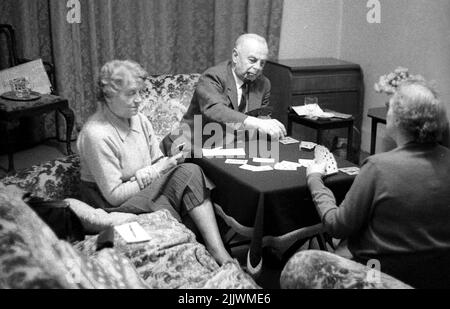 1960s, historisch, drei ältere Menschen, die in einer Lounge sitzen und Karten an einem kleinen Tisch spielen, England, Großbritannien. Der Mann trägt Anzug und Krawatte und raucht eine Pfeife. Stockfoto