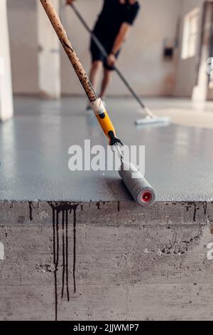 Eine vertikale Nahaufnahme von zwei Bauarbeitern, die in einer Industriehalle graues Epoxidharz auftragen Stockfoto